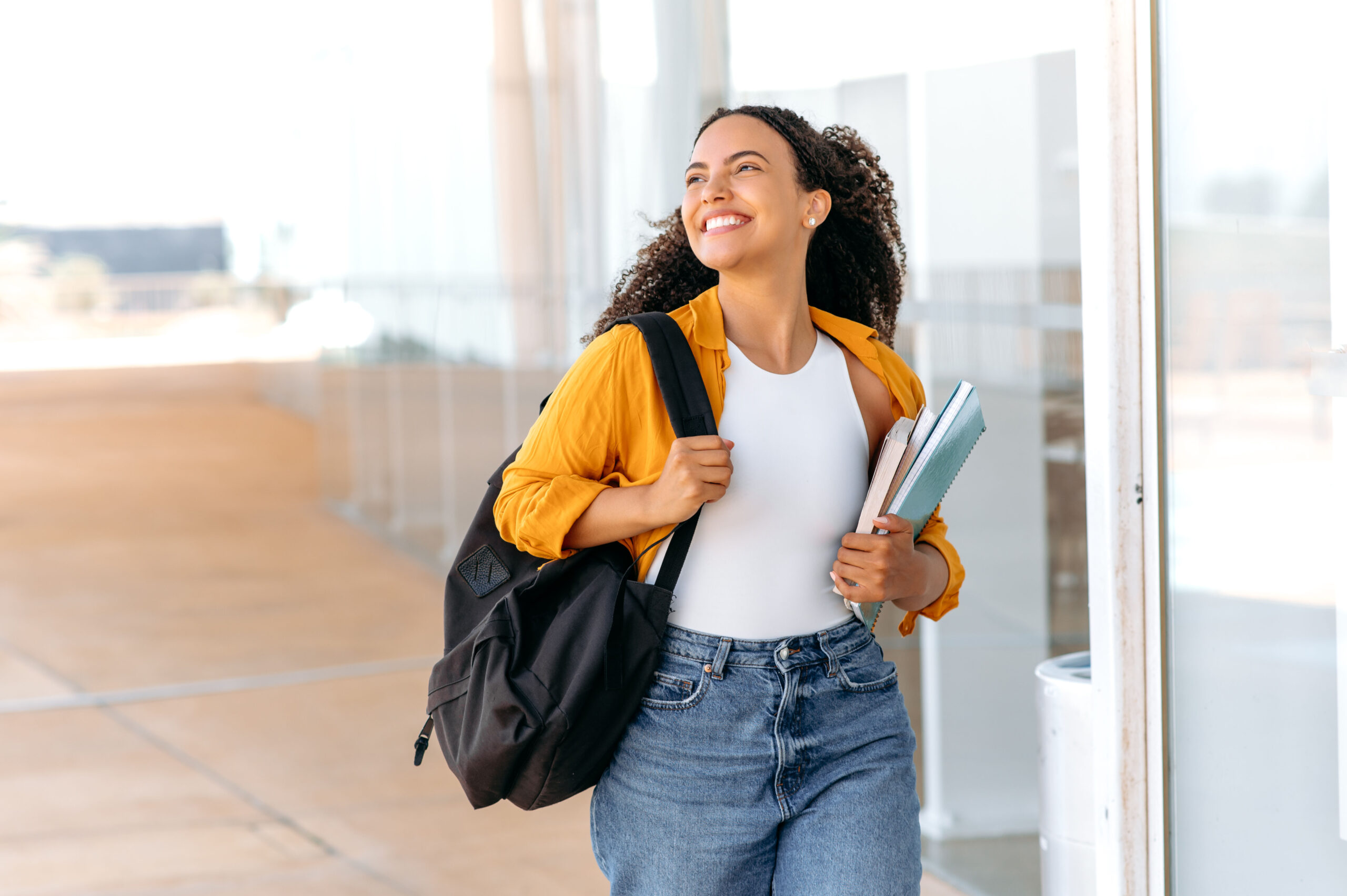 Happy lovely curly haired brazilian or hispanic female student, with a backpack, hold books and notebooks in her hand, walking near the university campus, looks away and smile, finished school day