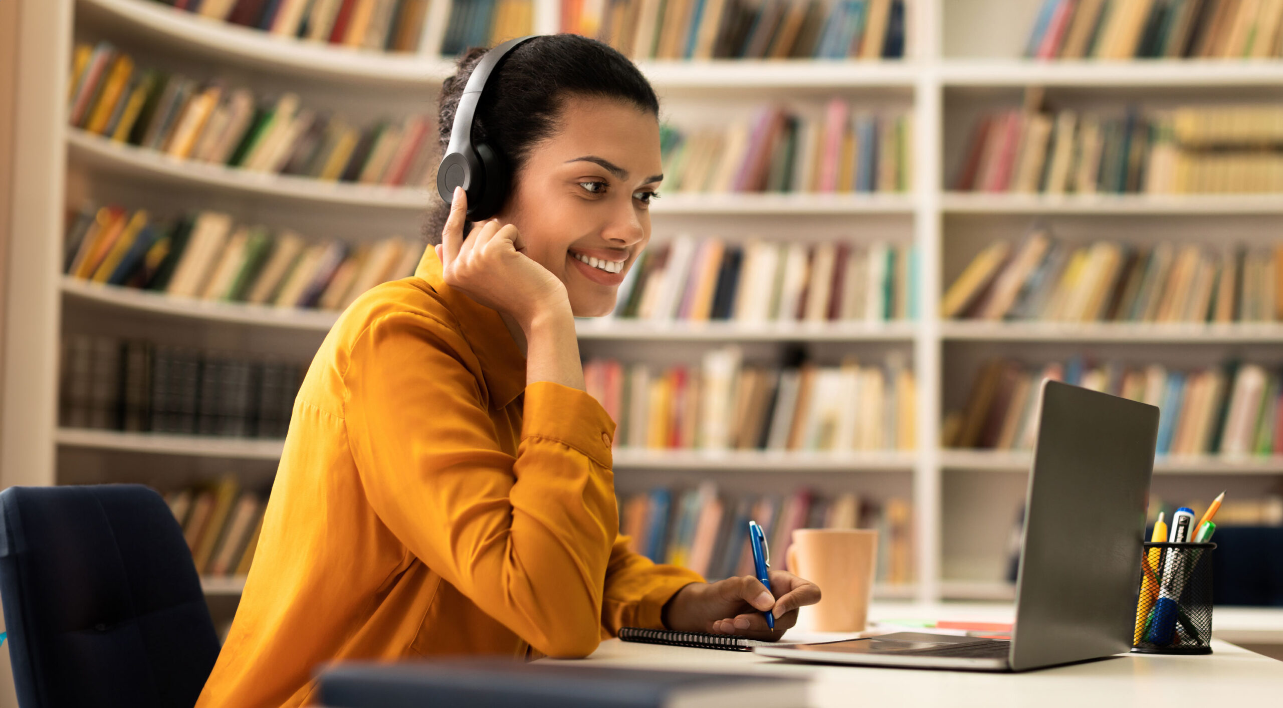 A female infront of a computer and using a headset. In the background it looks like shes in a library.