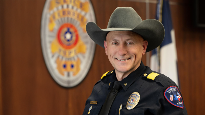 East Texas alum Arley Sansom, dressed in his police uniform and wearing his new chief's badge, smiles for a professional photo against a woodgrain wall featuring a large, out-of-focus Terrell Police Department seal.