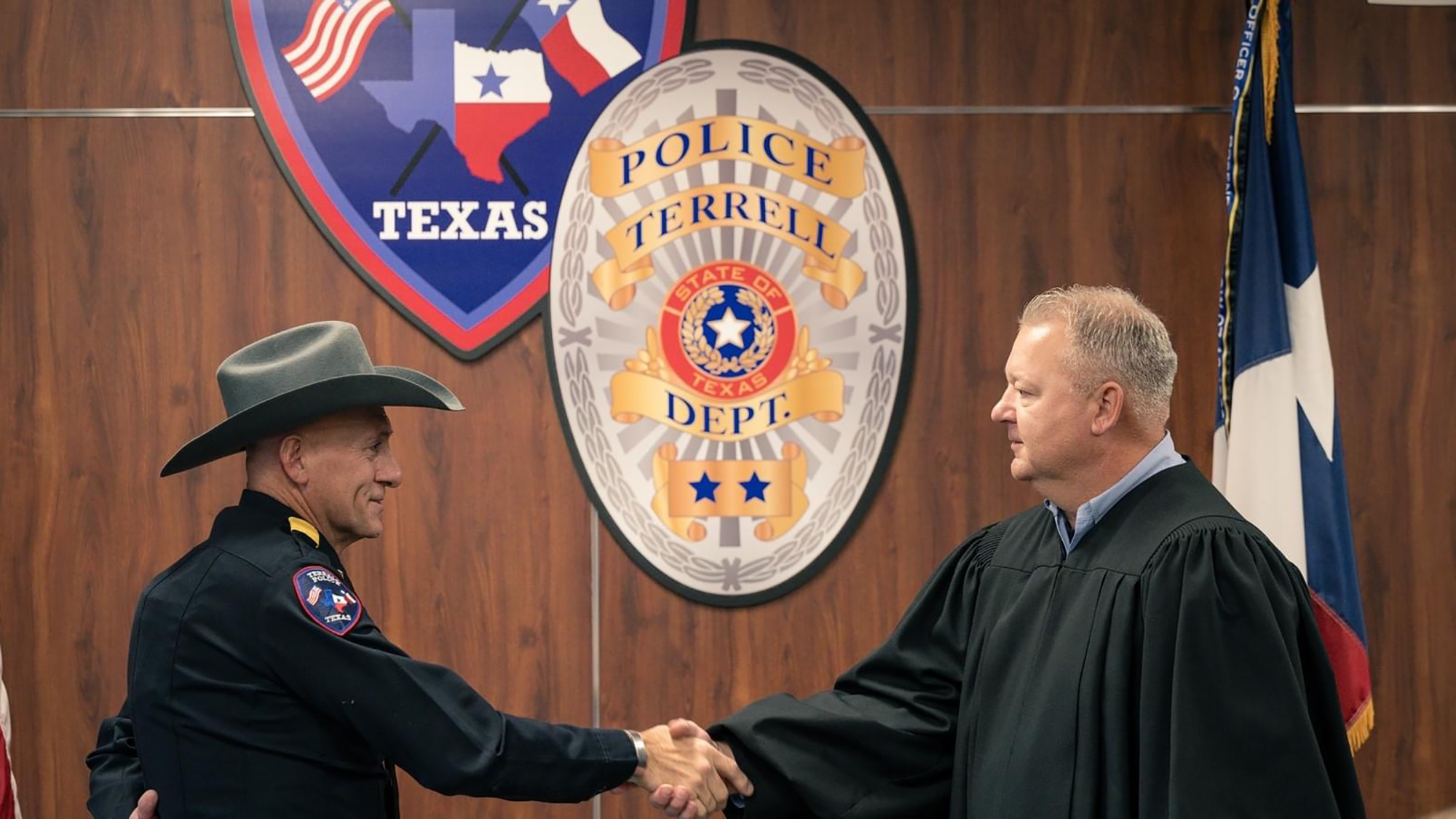 East Texas A&M alum Arley Sansom, dressed in police attire and a cowboy hat, shaking hands with Municipal Judge Mike Smith in front of a woodgrain wall featuring a large Terrell Police Department seal.