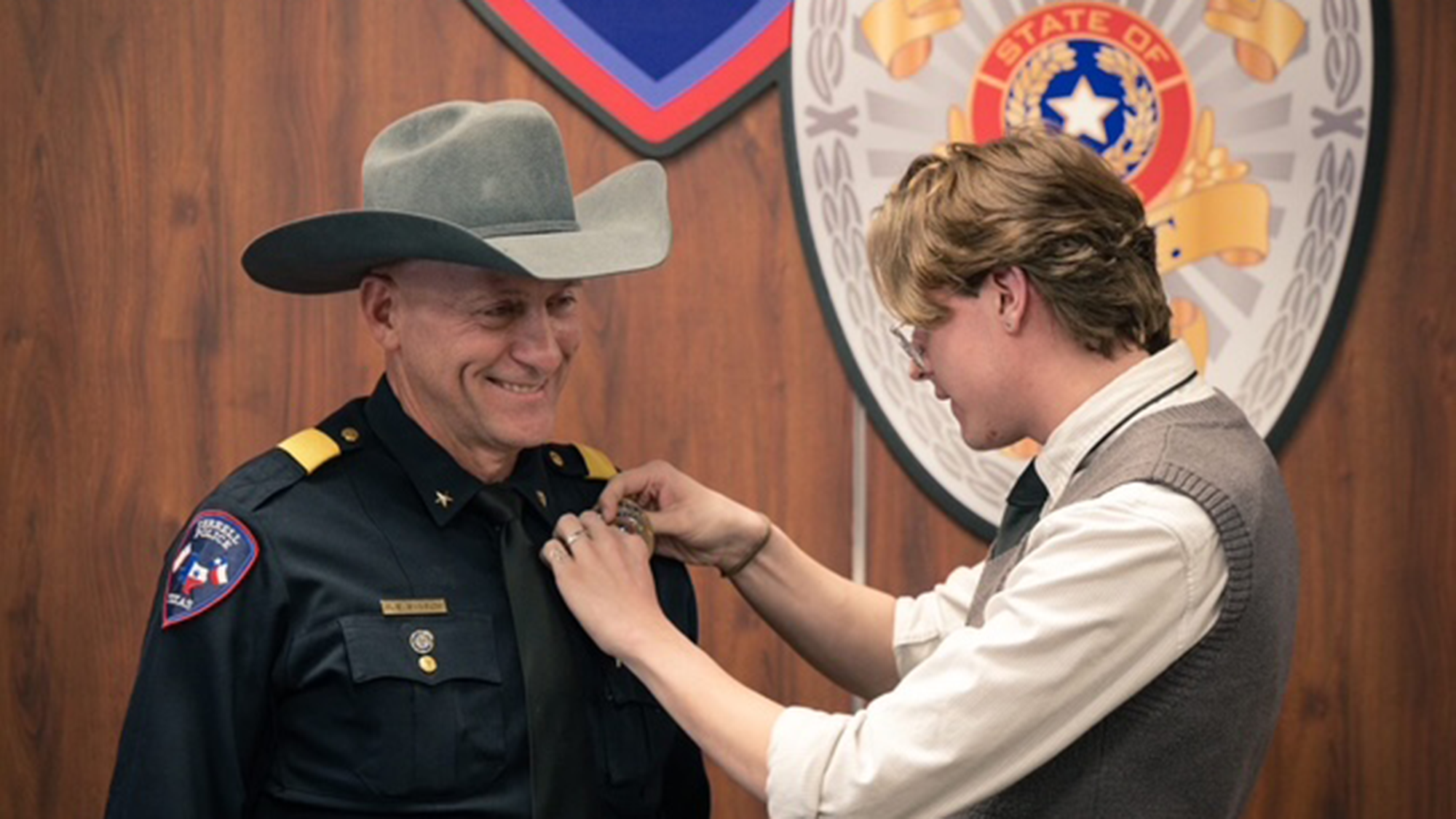 Cole Sansom pinning a badge on his father, Arley Sansom's uniform while both are standing in front of a woodgrain wall featuring a large Terrell Police Department seal.