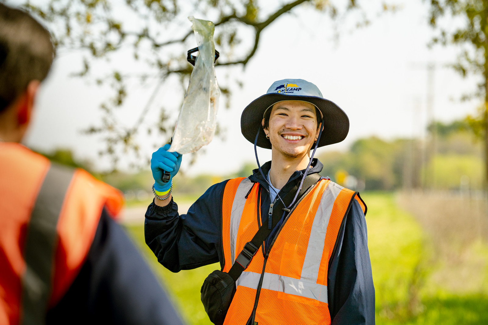 A smiling male holding a plastic bag. He is wearing a safety jacket and a hat. There is someone else in front of him with the same outfit and their outside.