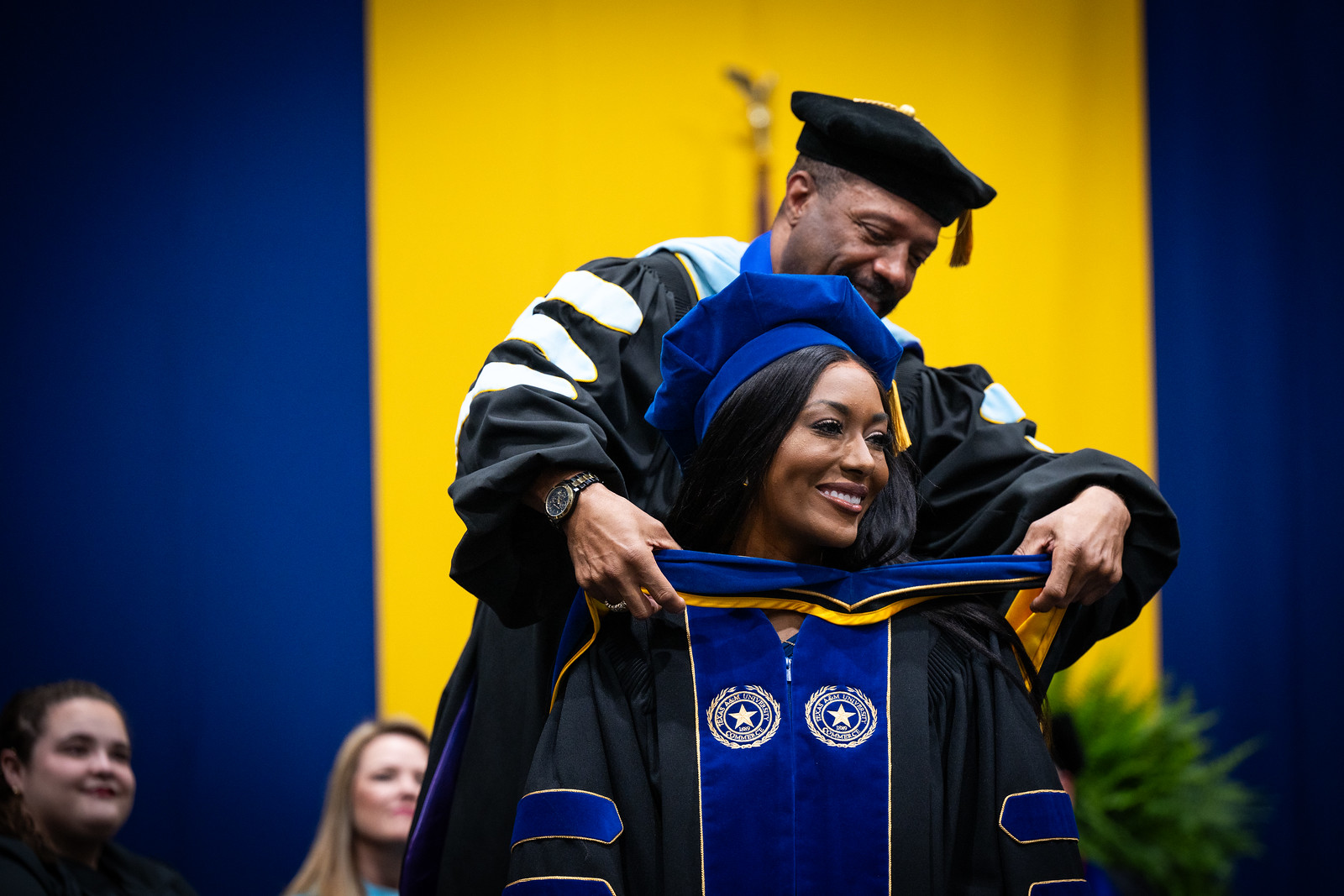 A female graduate is receiving a scarf from a professor. In the background is yellow and blue with two other people facing foreward.