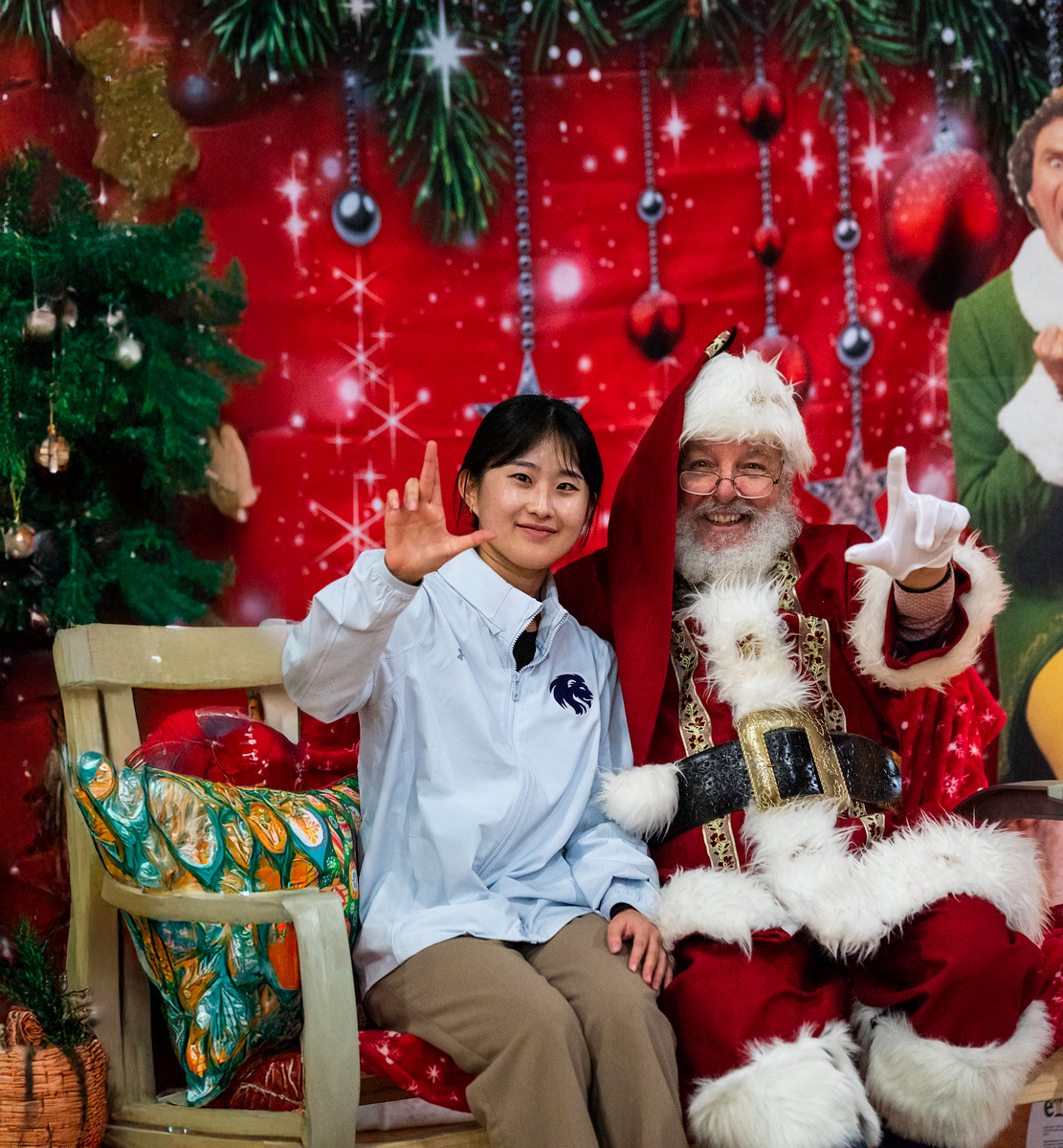 A person in a light blue jacket sits with Santa Claus in a festive holiday setting, featuring red ornaments, greenery, and sparkling decorations.