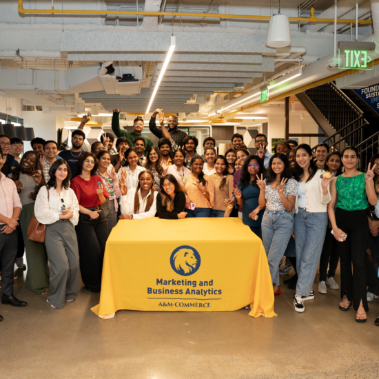 A large group of people pose for a photo around a table covered with a cloth featuring the logo for the department of marketing and business analytics at A&M-Commerce.