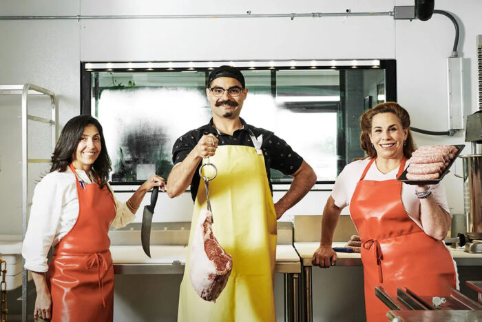 Three butchers stand in a kitchen holding cuts of meat.