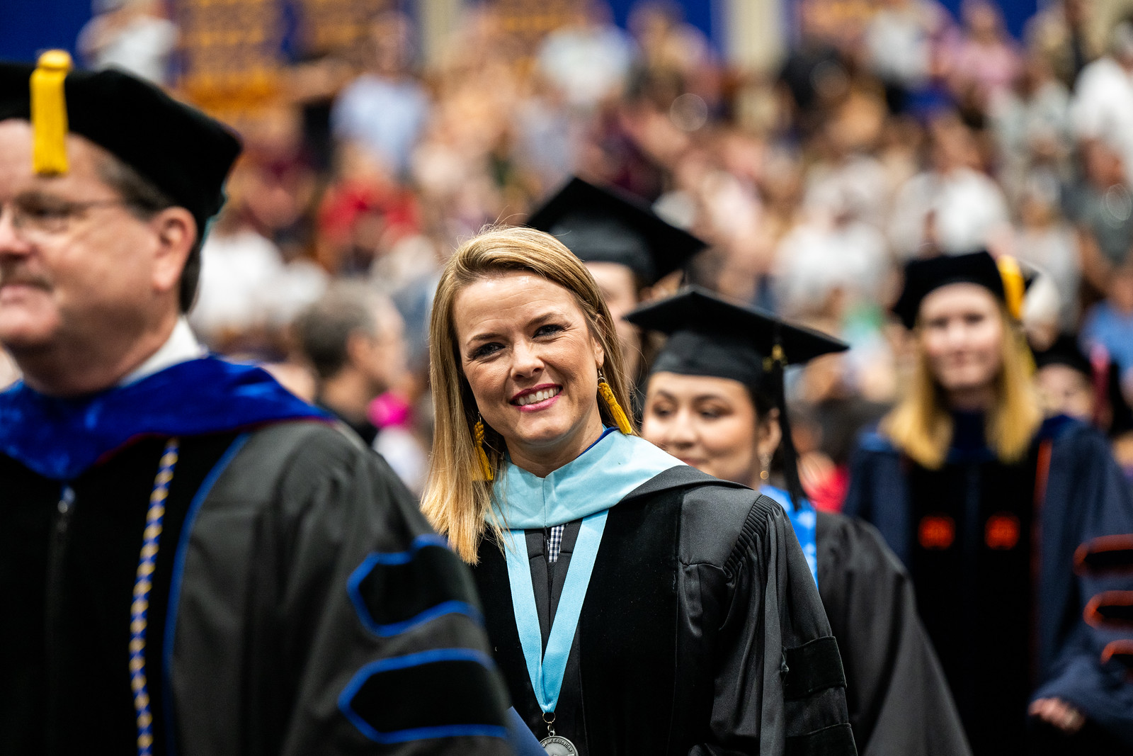 A female graduate student smiling at the camera man. She appears to be in a line with other graduates and in the background seems to be the audience.