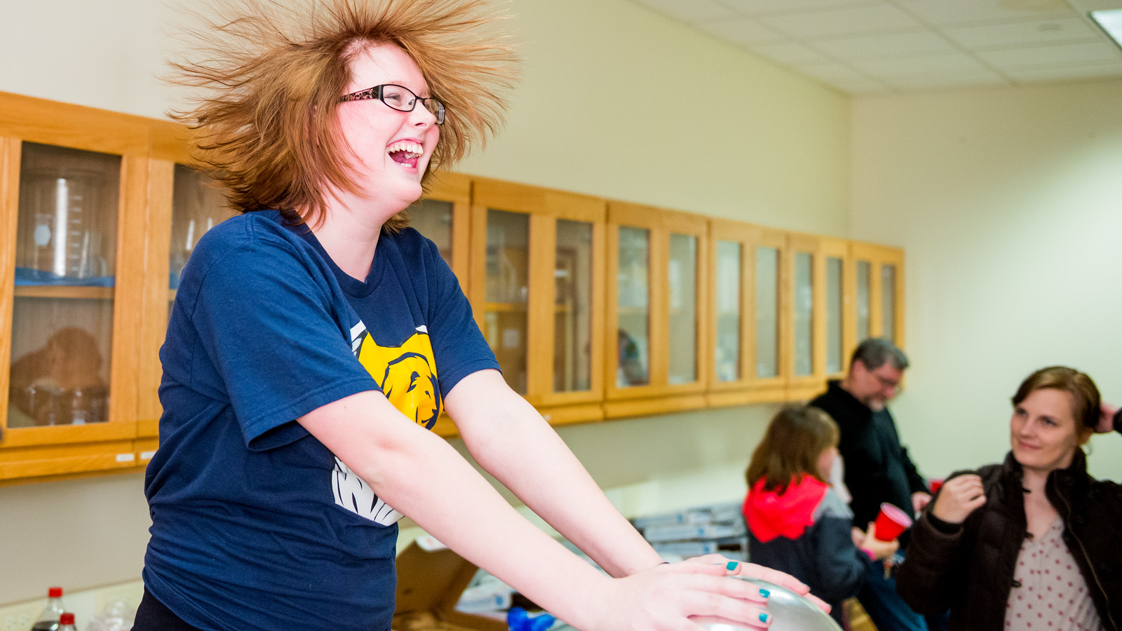 A student touches a van de graaff generator, resulting in their long, red hair standing on end.