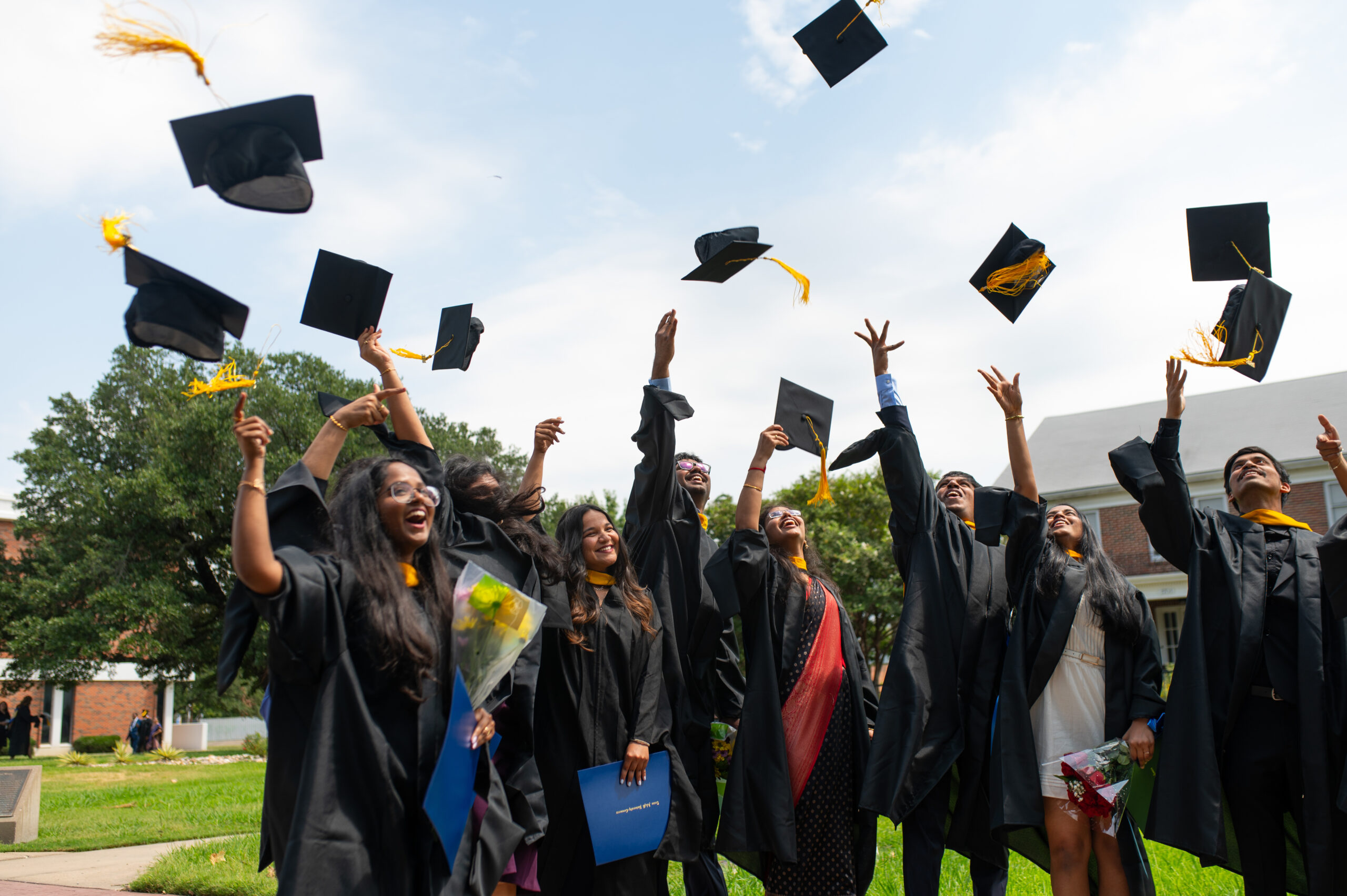 Graduates in black gowns toss their caps into the air. Blue sky with clouds and green grass are in the background.