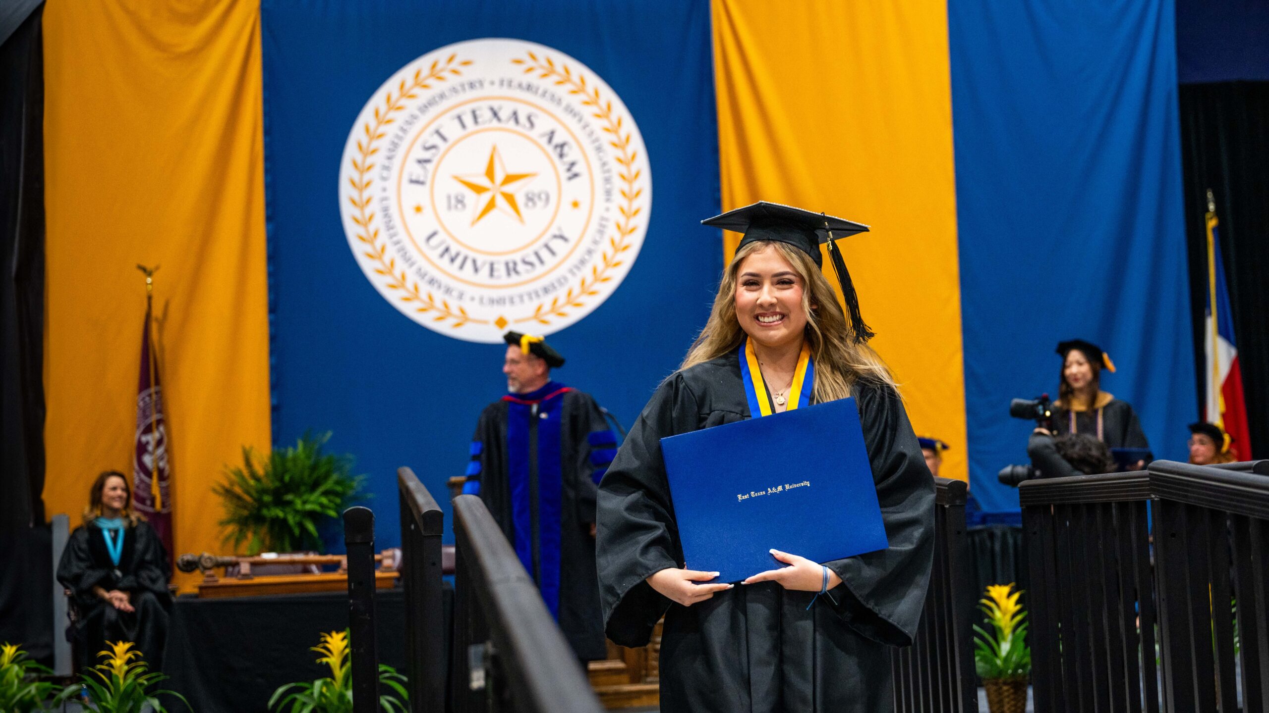 Student smiles with diploma in hand and graduation cap on her head. The university's seal and the graduation stage are behind her.