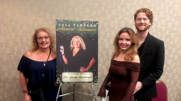 At left, Dr. Sandy Kimbrough stands next to a 2024 TAHPERD Award Winners sign showing a picture of her. On the right side of the image stands a female student with a male student behind her.