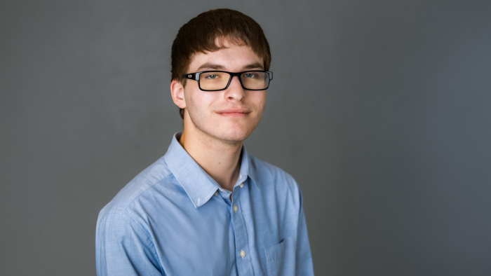 East Texas A&M cybersecurity alum Tag Kalat smiles at the viewer, wearing black-rimmed glasses and a light blue button-up against a gray background.