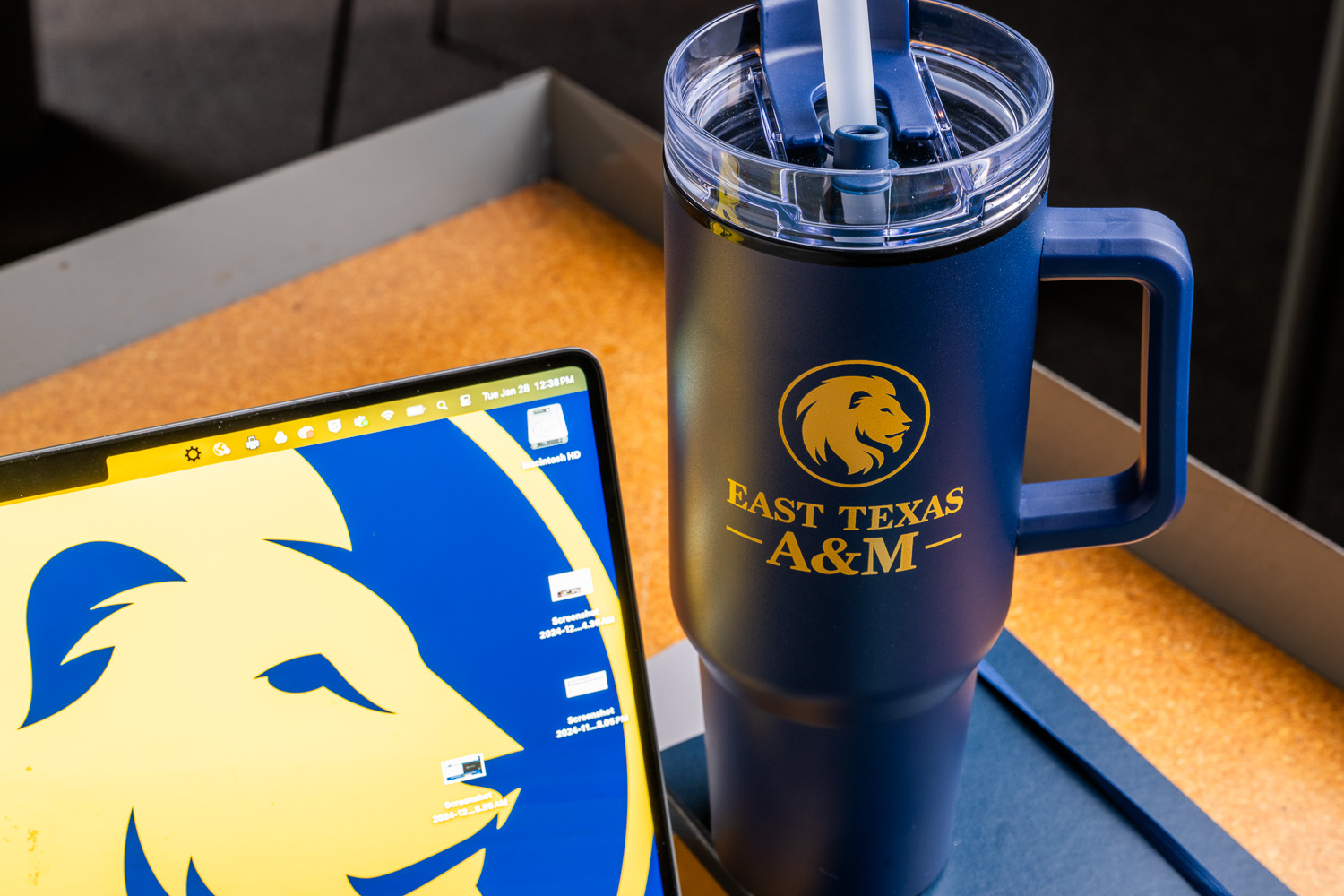 Blue tumbler with East Texas A&M logo and laptop featuring matching lion emblem on screen, photographed on a desk.