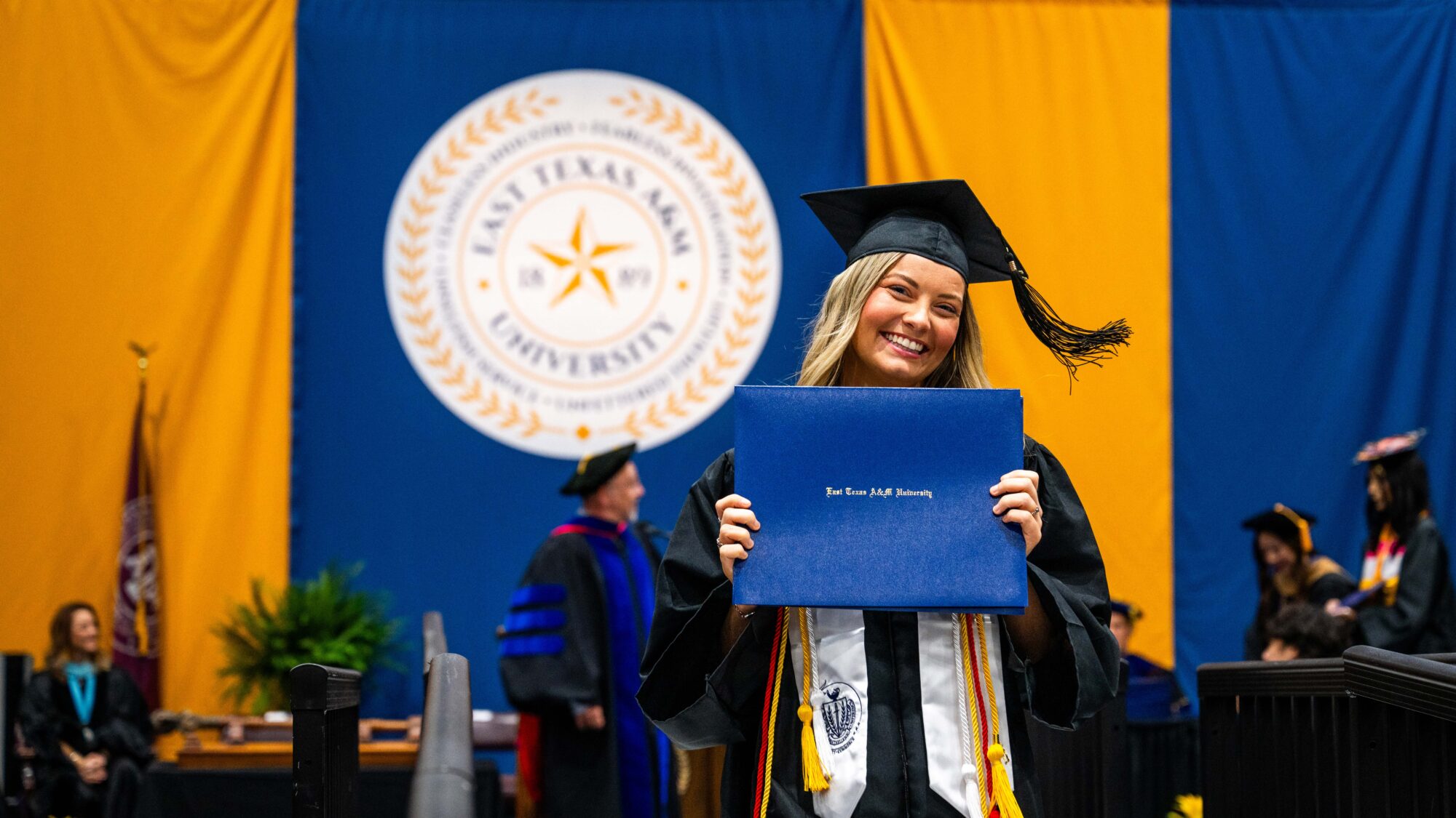 Graduate smiles with graduation cap on her head and holding blue diploma cover in front of her in both hands. The round university seal is behind her.