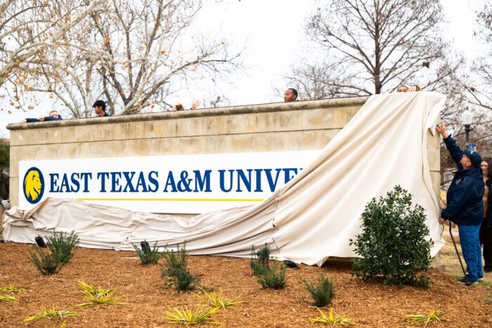 Several people stand behind the sign and release the canvas covering over the sign. A man on the far right reaches up to finish removing the canvas covering.