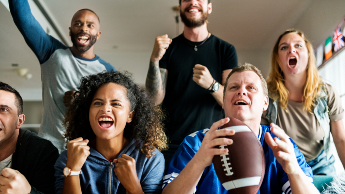 Six male and female football fans cheer their team on at a home watch party.