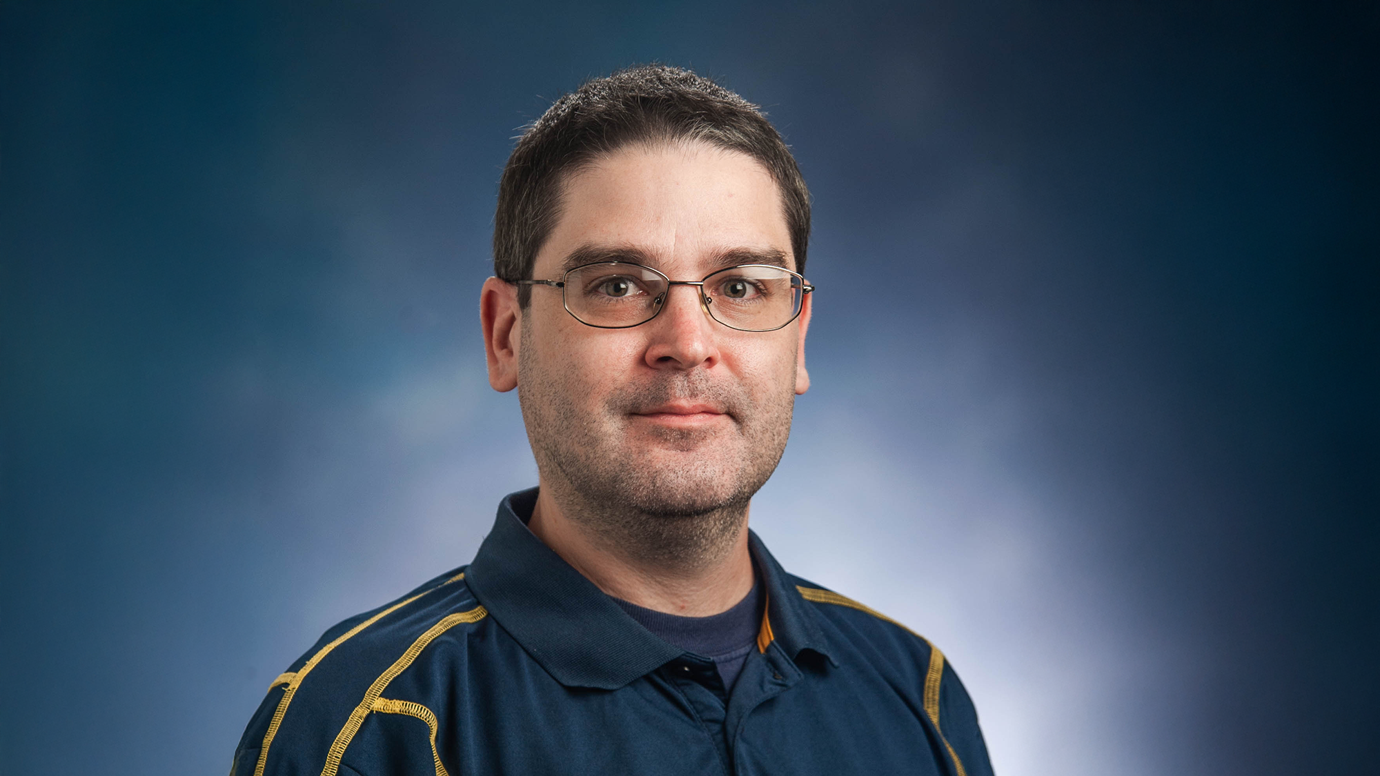 Professional headshot of Dr. Stephen Reysen against a mottled blue background, wearing a blue polo with yellow seams.