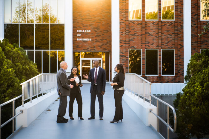 Four people standing outside of a brick building and conversing. The building features a sign that reads "College of Business."