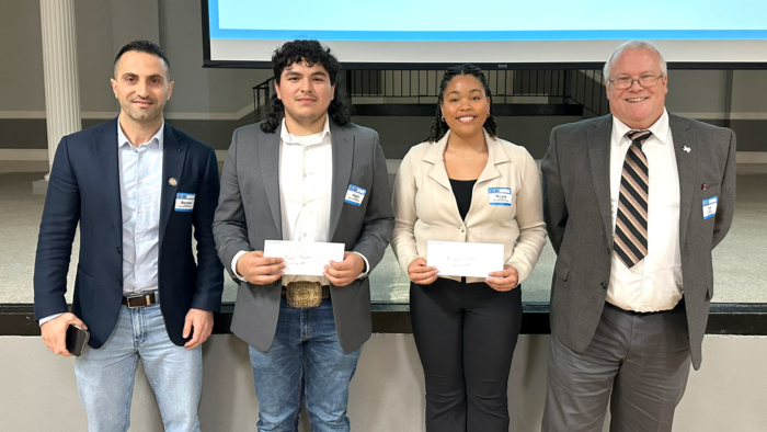 Four people stand shoulder to shoulder for a photo opportunity. The two people in the middle are students holding their scholarship envelopes.